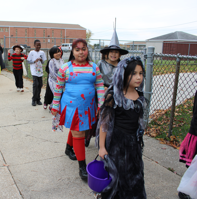 Children in Halloween costumes walk along a school sidewalk beside a chain-link fence, including a witch and a girl in a blood-spattered blue dress; festive mood.