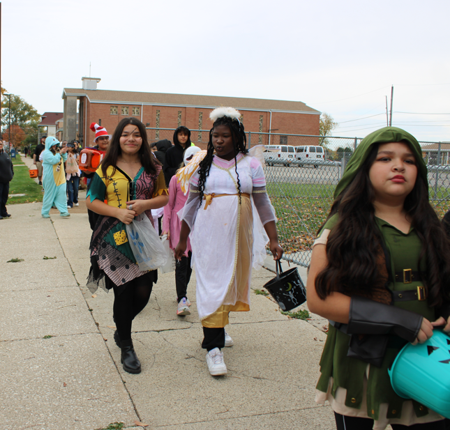 Students in colorful Halloween costumes walk along a school sidewalk; a girl in a white gown and another in a green hood lead the group, with a festive, relaxed mood.