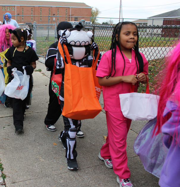 Children in Halloween costumes walk along a sidewalk carrying treat bags; a girl in pink scrubs leads beside a child in a cartoon dog mask, creating a lively, festive mood.