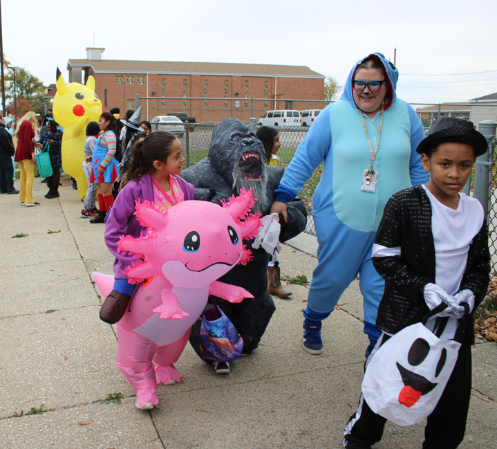 Children in colorful Halloween costumes parade along a school walkway, carrying an oversized pink axolotl and a ghost bag as a smiling adult chaperone walks with them.