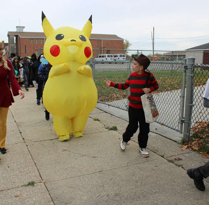 A child in a red-and-black striped shirt greets a person in a large Pikachu costume on a school sidewalk, as classmates look on; playful, festive mood.