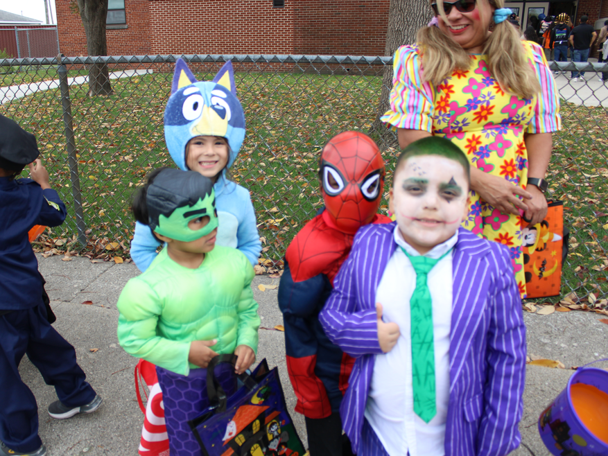 Children dressed as Bluey, Hulk, Spider-Man and the Joker pose with a smiling adult in a bright floral outfit on a school sidewalk, in a cheerful Halloween scene.