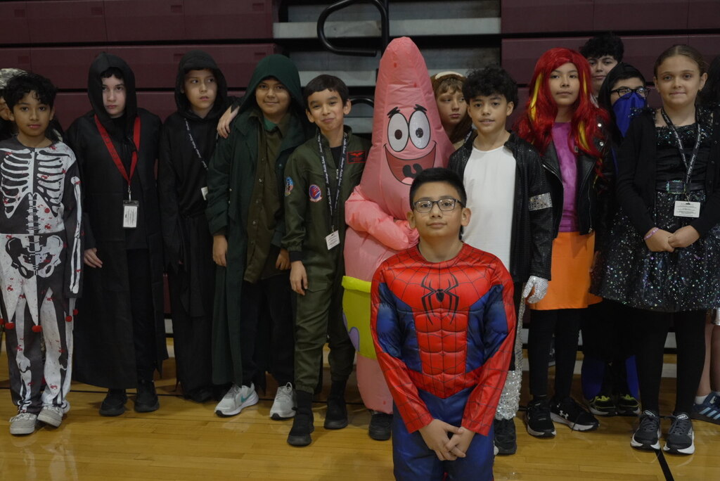 Children in Halloween costumes pose in a school gym, with Spider-Man kneeling in front and Patrick Star, skeletons, and superheroes behind him.