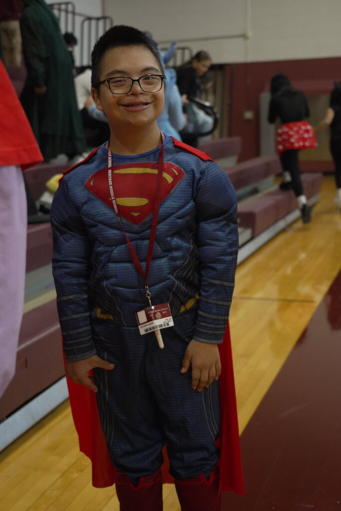 Smiling child in glasses wears a padded Superman costume, red cape, and lanyard, posing on a school gym floor as people move in the background.