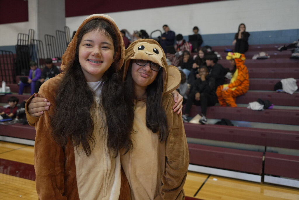 Two smiling students in animal onesie costumes pose arm in arm in a school gym, with classmates in costumes on the bleachers behind them.