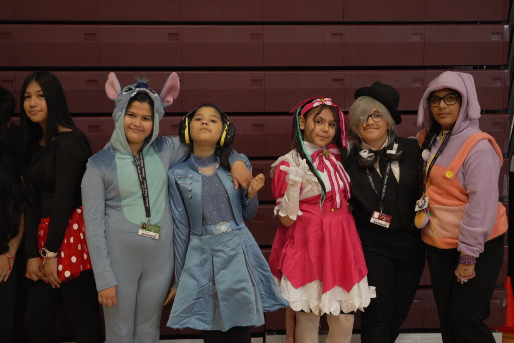 Six young women in colorful cosplay costumes pose together before maroon bleachers, wearing animal ears, headphones, wigs, and playful outfits at a convention.