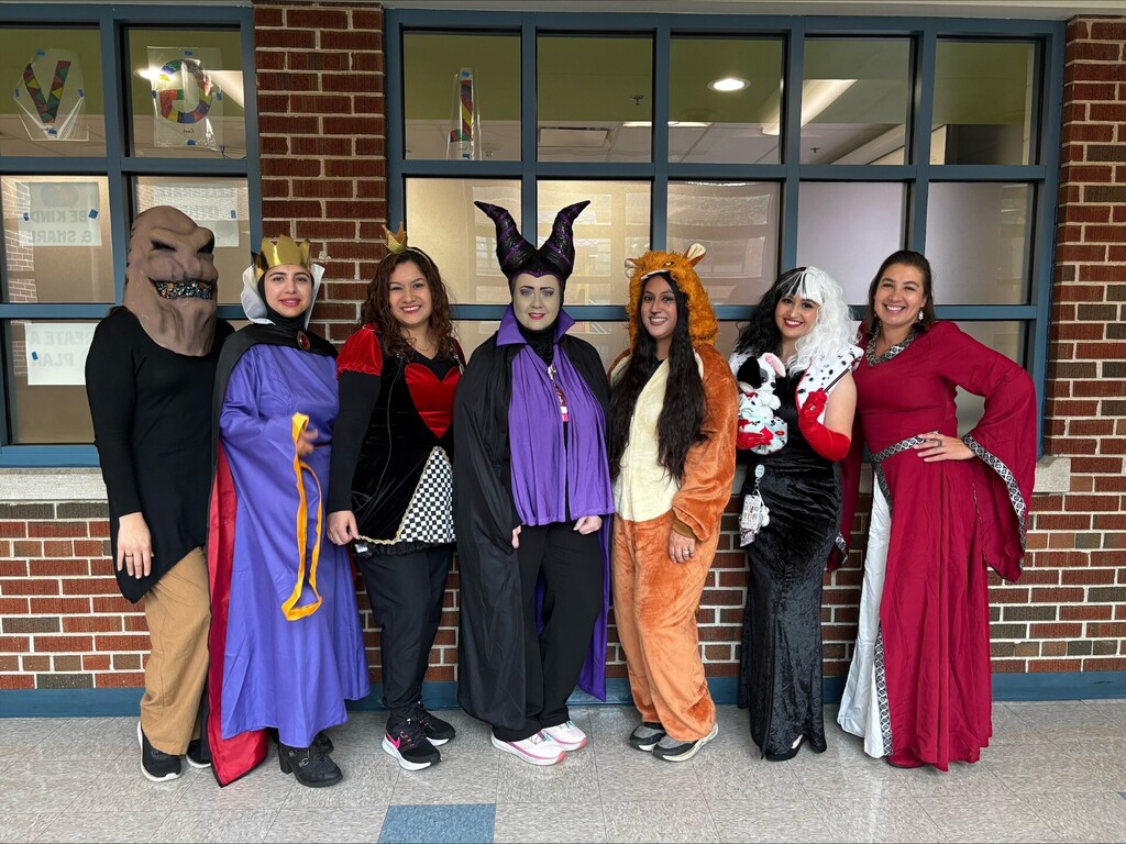 Seven adults pose in Halloween costumes, including witches, animal suits and a red gown, smiling in front of brick school windows.