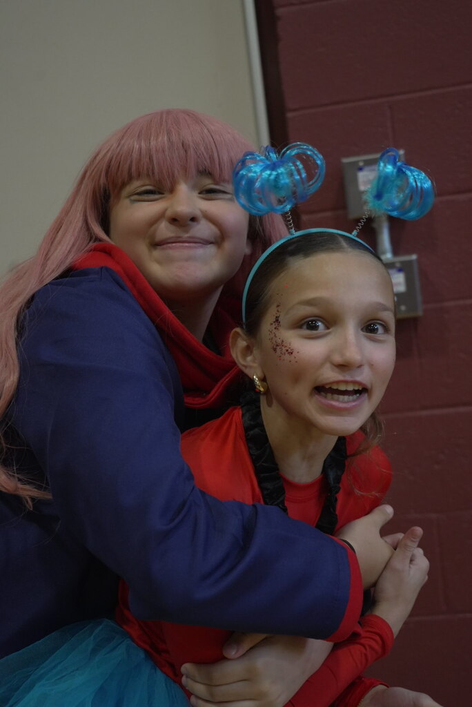 A smiling woman in a pink wig hugs a grinning girl in a red-and-blue costume and turquoise skirt, wearing a playful blue antenna headband.