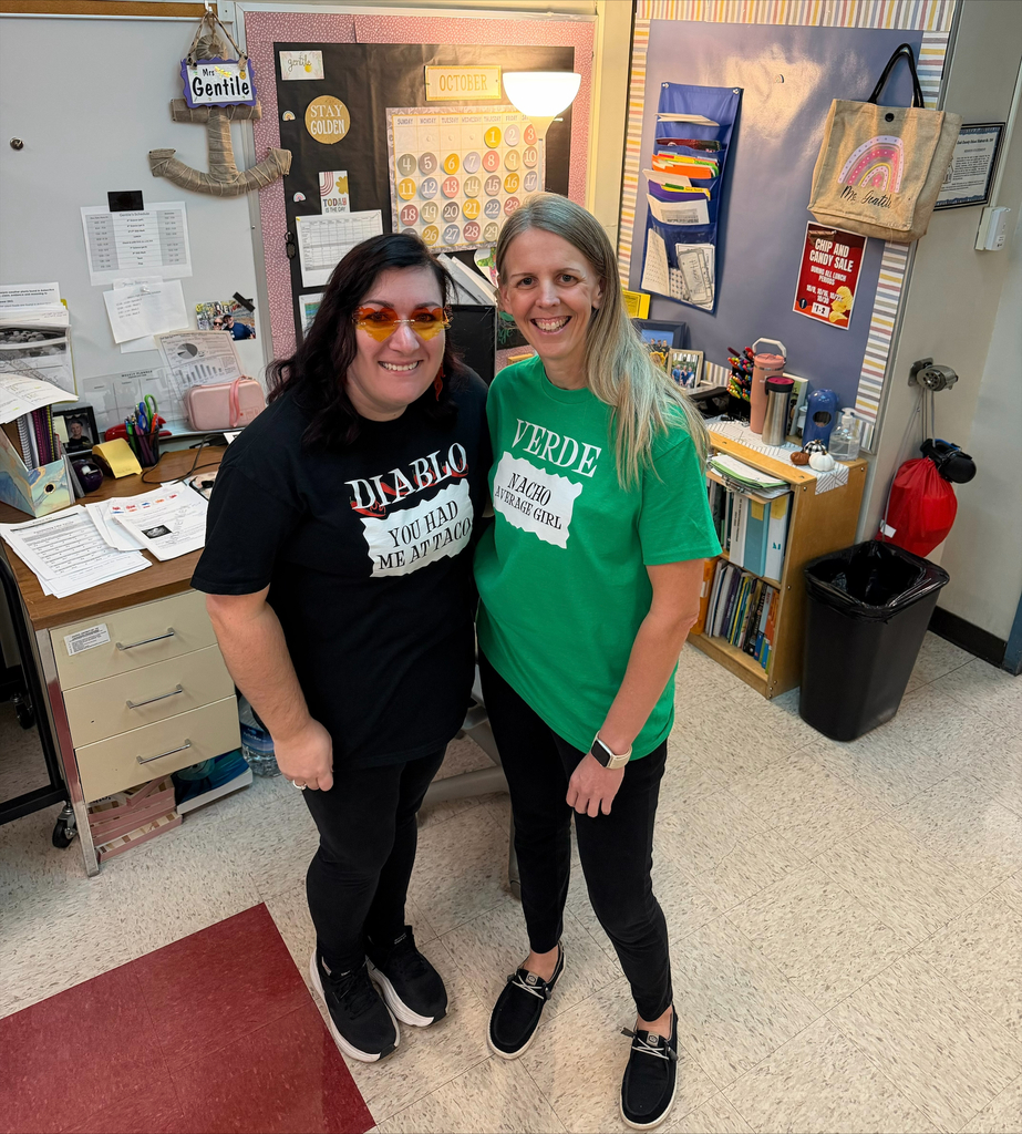 Two smiling women stand arm in arm in a colorful classroom, surrounded by student artwork, books, desks, and teaching materials, conveying warmth and camaraderie.