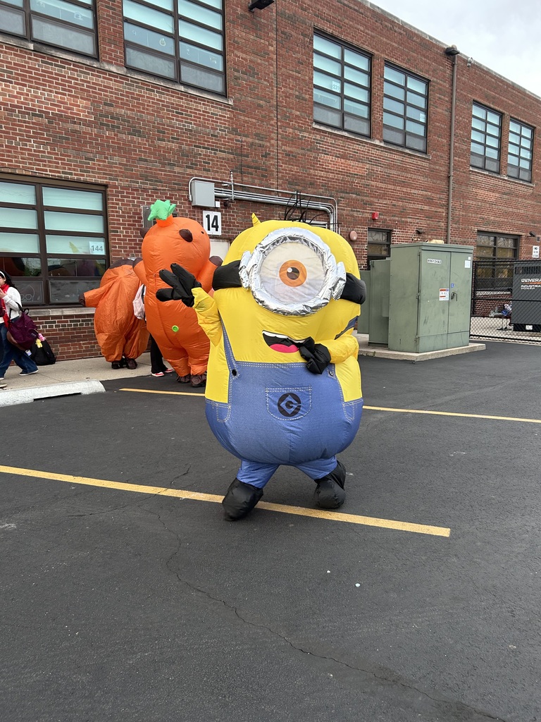 A cheerful one-eyed Minion in blue overalls poses beside an oversized orange carrot mascot in a school parking lot, with a brick building behind them.