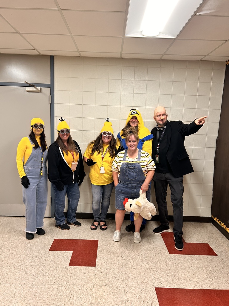 Six adults pose in a tiled hallway, several dressed as Minions in yellow and denim; a man points while a woman holds a plush toy. Playful mood.