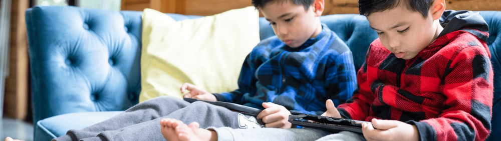 two young boys sit side by side looking at their own tablet devices
