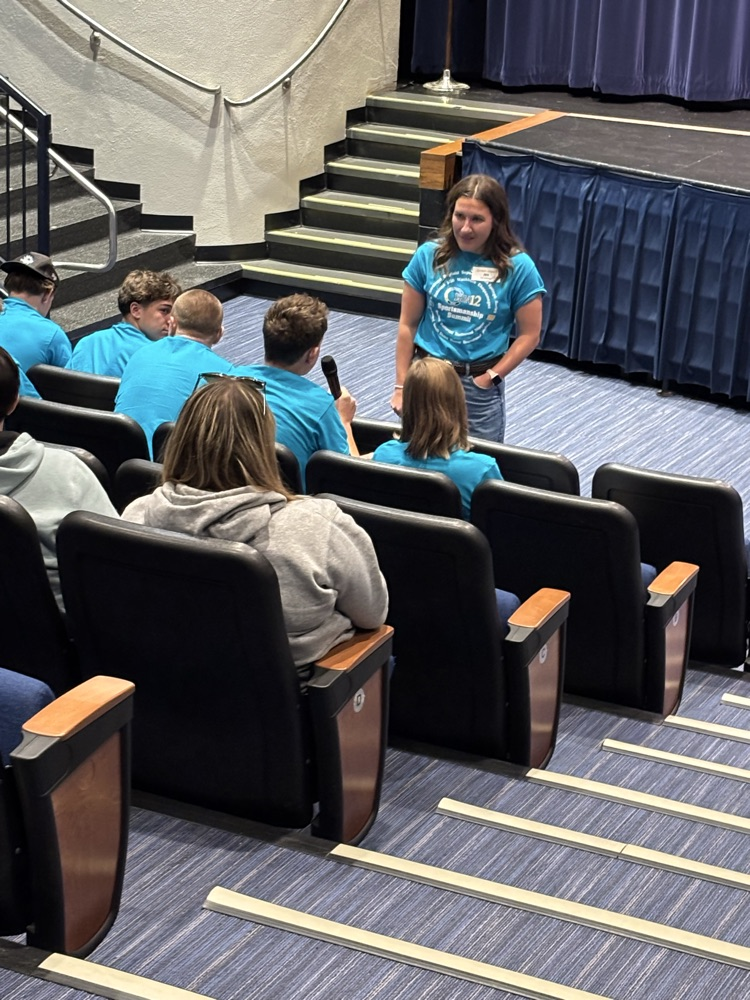 Presenter Ivie Brooks is standing by the chairs the audience is sitting in and has handed the microphone to a student to share their answer to a question. 