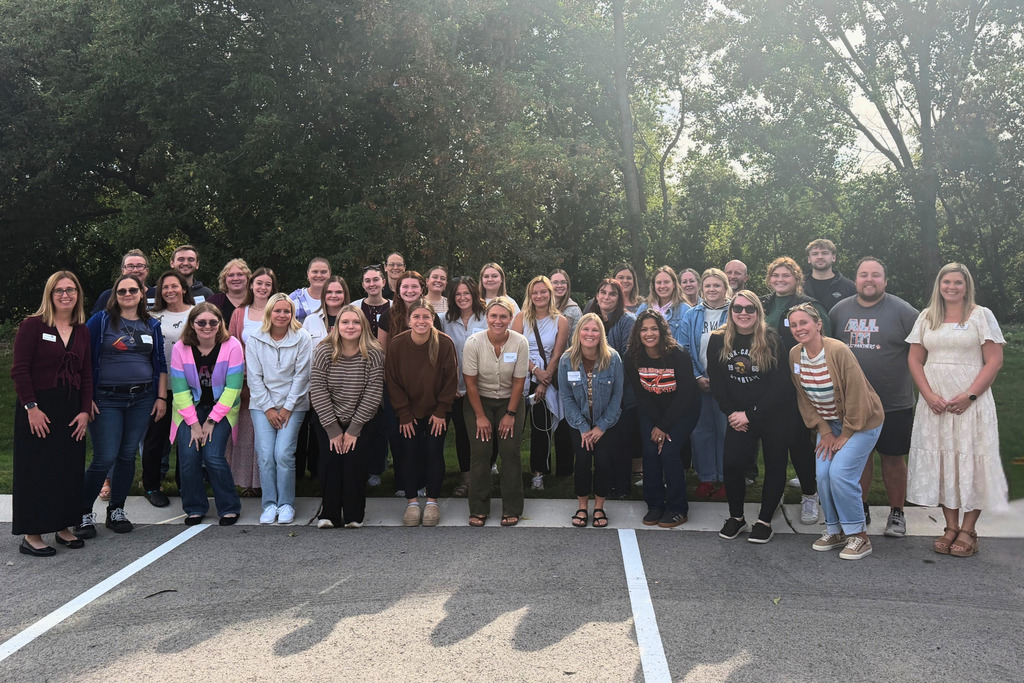 A group of people in various outfits poses for a photo in a parking lot with trees.
