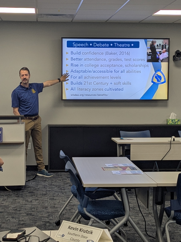 A man presenting in a classroom, standing near a screen displaying benefits of speech and debate.