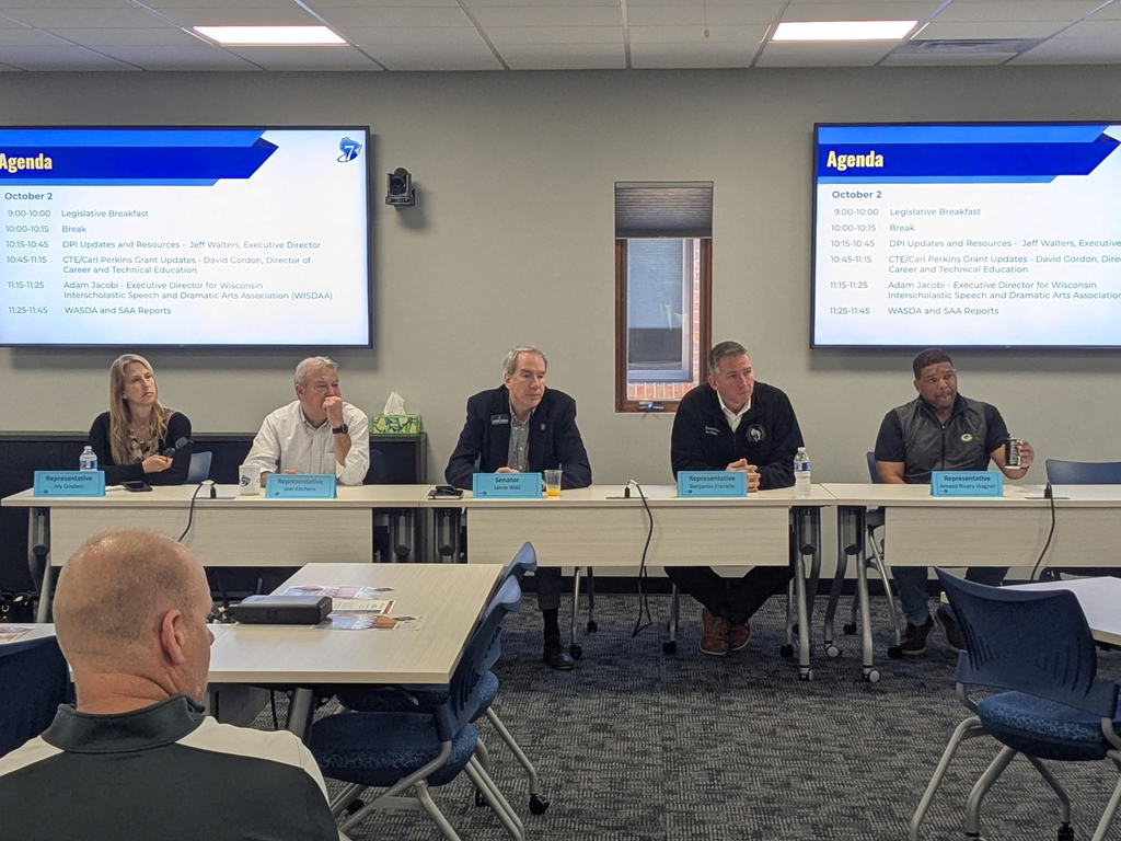 Five people sit at a table in a meeting room, with two screens displaying text behind them.