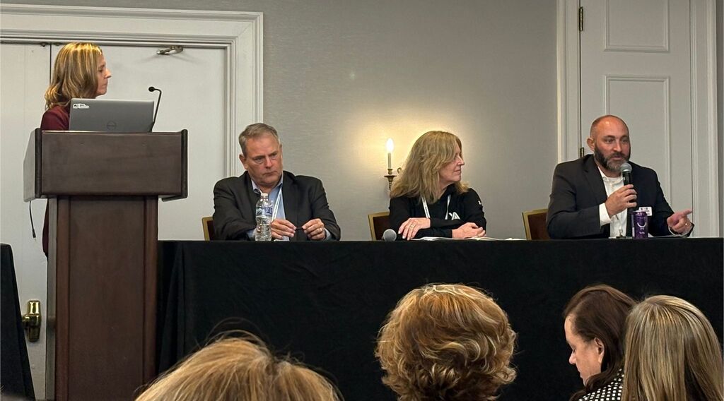 A group of four seated people behind a table, one person at a podium, and several people in front.