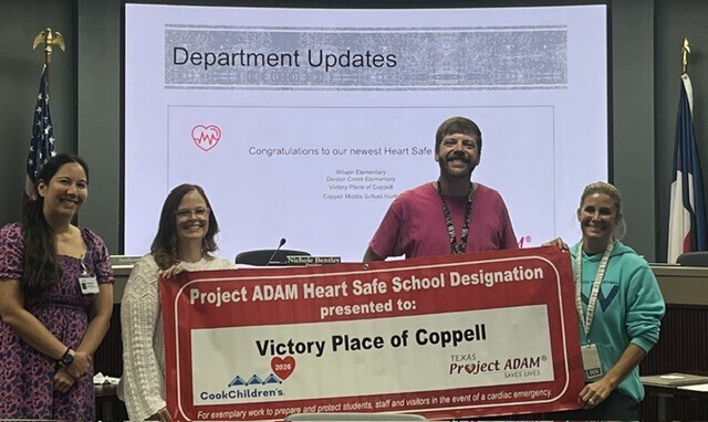 Four adults in a meeting room hold a banner recognizing Victory Place of Coppell as a Project ADAM Heart Safe School.