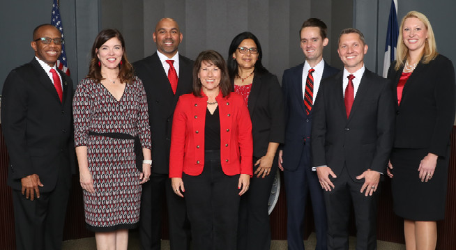 Group photo of eight Coppell ISD board of trustees and the superintendent, dressed in business attire, smiling together in front of a stage backdrop with American and Texas flags..