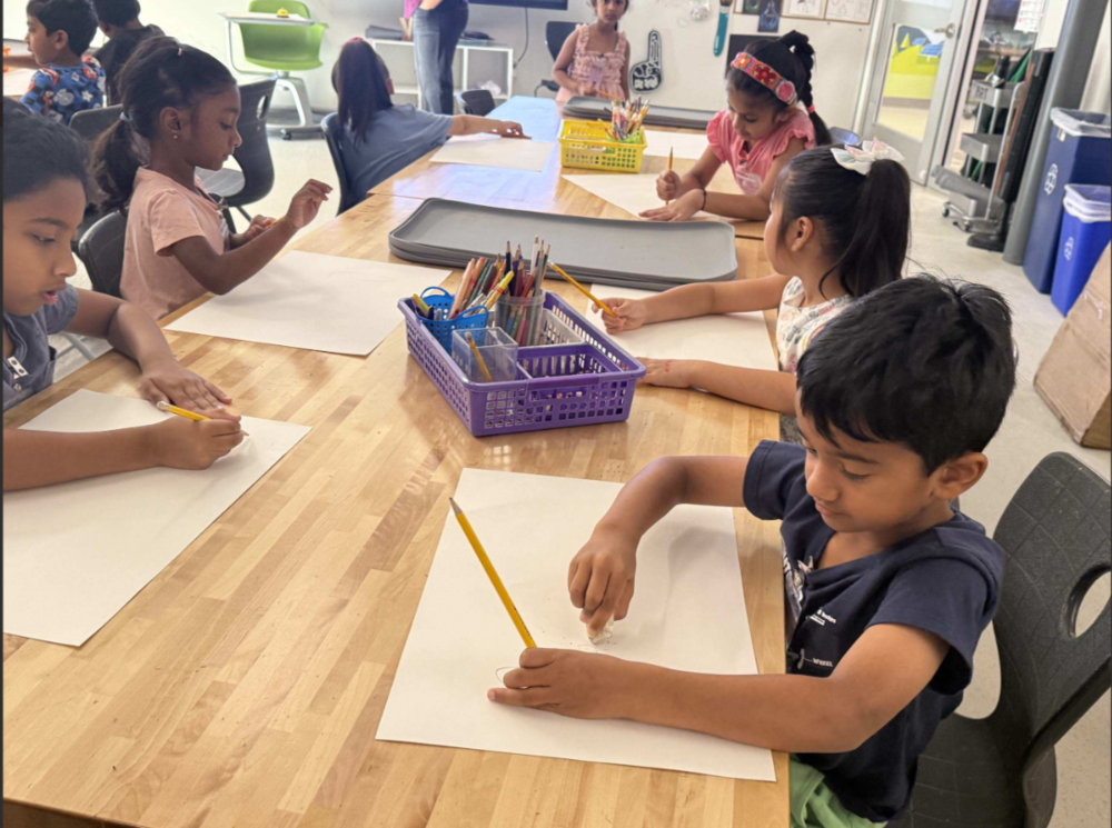 Students sit around a classroom table using pencils and paper during an art or writing activity, with baskets of colored pencils and supplies in the center.