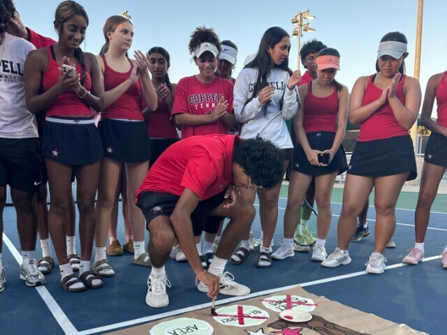 Coppell tennis players gather on an outdoor court and applaud as a man in a red shirt paints an x over circles on a banner.