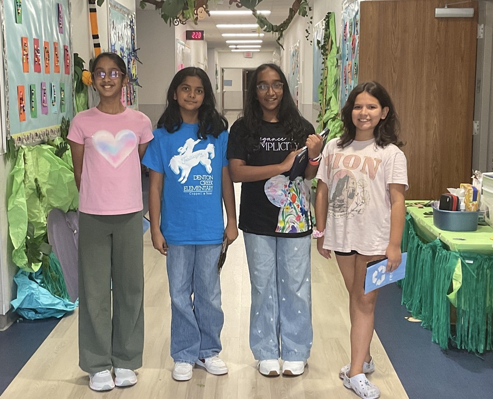 Four students stand in a school hallway decorated with green, nature-themed displays and hold recycling materials after participating in a classroom recycling activity.