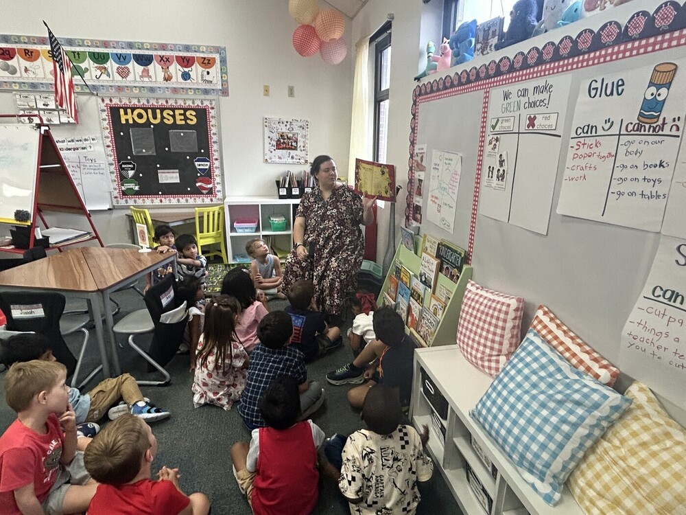 Madison Smith reads a book to a group of young students seated on a classroom rug. The classroom includes alphabet cards, picture books, learning posters and anchor charts with examples of classroom choices and how to use glue.