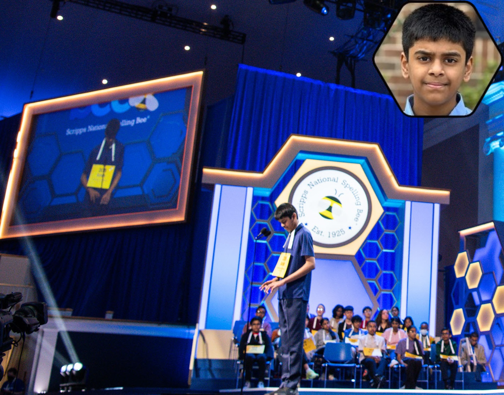 Krish Manjunath stands on a stage at the Scripps National Spelling Bee, facing a microphone while wearing a number tag on a lanyard. Behind Krish Manjunath, other contestants sit in rows of chairs. The stage features a large honeycomb-themed backdrop with the “Scripps National Spelling Bee” logo, bright blue lighting, and a large screen on the left displaying the student. A small inset image in the upper right shows a close-up headshot of Krish Manjunath.