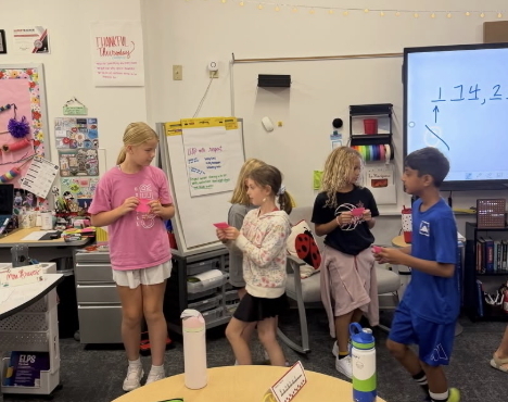 Students stand in a classroom holding cards during a math activity near a whiteboard, classroom materials and an interactive display.