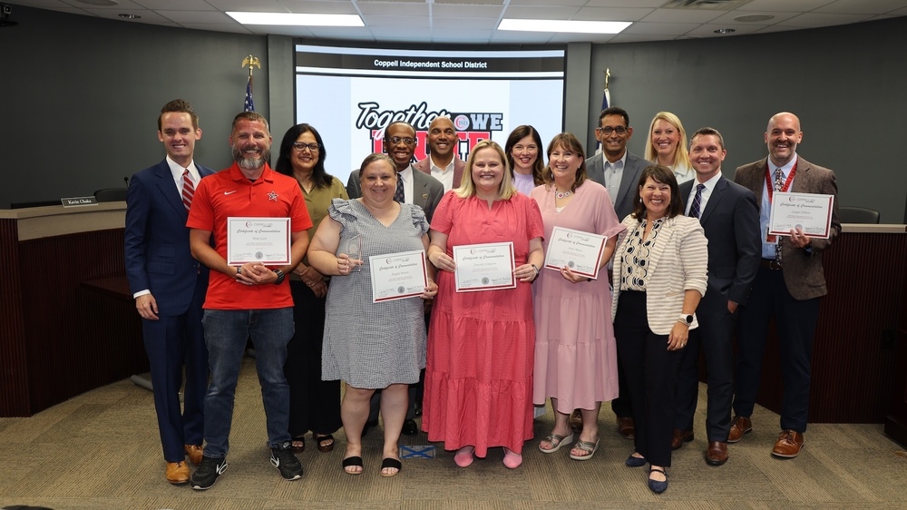 Group photo in the Coppell ISD board room showing 14 staff members and administrators standing in front of a presentation screen displaying the message “Together We Unite” beneath the heading “Coppell Independent School District.” Five individuals in the front row hold Certificates of Commendation. A U.S. flag and Texas flag are visible on either side of the screen. The group is posed for recognition, with board room seating and nameplates visible in the background.