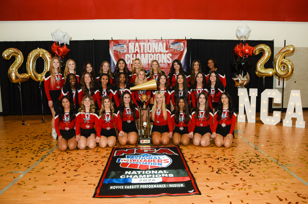 Group photo of the Coppell Cheer team posing in matching red-and-black uniforms inside a gym, arranged in rows around a large championship trophy. Behind them is a banner reading “National Champions,” with black, red, and gold balloons shaped like “2026” and illuminated “NCA” letters. The floor mat in front reads “NCA National Cheerleaders Association National Champions 2026 – Novice Varsity Performance Medium,” with gold confetti scattered across the floor.