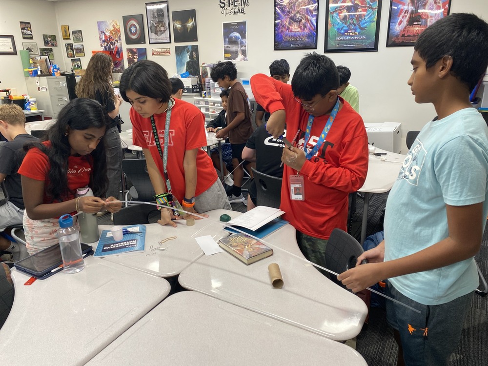Students work in small groups on a hands-on classroom activity using straws, paper, tape and other materials at desks.
