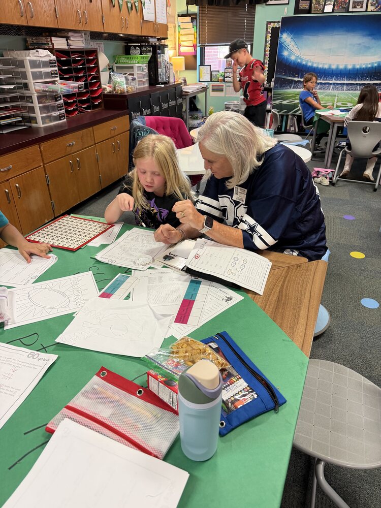 A teacher wearing a Dallas Cowboys jersey helps a student complete a worksheet at a classroom table while other students work in the background.