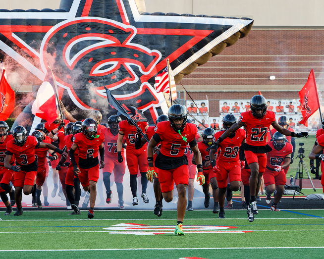 Football players in red uniforms run onto the field through smoke and a large team mascot tunnel before a game, with flags waving behind them.