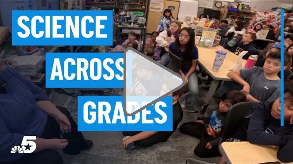 A classroom filled with elementary students seated at tables and on the floor, listening to an adult leading a lesson. Bold text on the left reads “Science Across Grades,” with a play icon over the image, suggesting a video about hands-on science learning in a collaborative classroom setting.