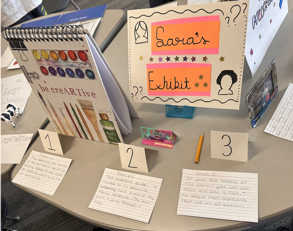 A student-created “Sara’s Exhibit” display is arranged on a classroom table. The center sign reads “Sara’s Exhibit” on orange and pink paper, decorated with stars, question marks, and silhouette drawings. Three numbered items are displayed with handwritten description cards. Item 1 is a sketchbook featuring watercolor paints, colored pencils, and the phrase “be creARTive.” Item 2 is a pack of collectible trading cards. Item 3 is a yellow pencil. A small framed photograph is displayed to the right of the exhibit. Additional student projects and note cards are visible on surrounding tables in the classroom.
