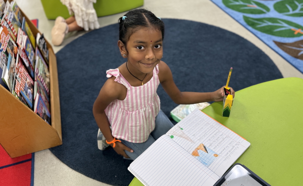Student sits at a green classroom table holding a pencil next to an open notebook with writing and a drawing. A book display, rug, tablet and classroom materials are visible nearby.
