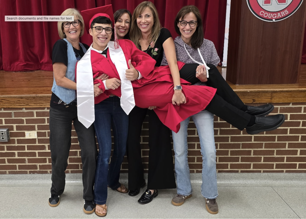 Five adults hold a graduate in a red cap and gown horizontally onstage as the graduate gives a thumbs-up.