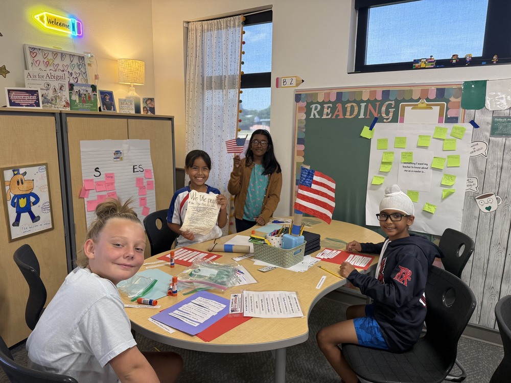 Students sit around a classroom table during a Constitution Day activity, holding U.S. flag projects, “We the People” materials and worksheets about the U.S. Constitution. The classroom includes a reading board, student work and patriotic decorations.