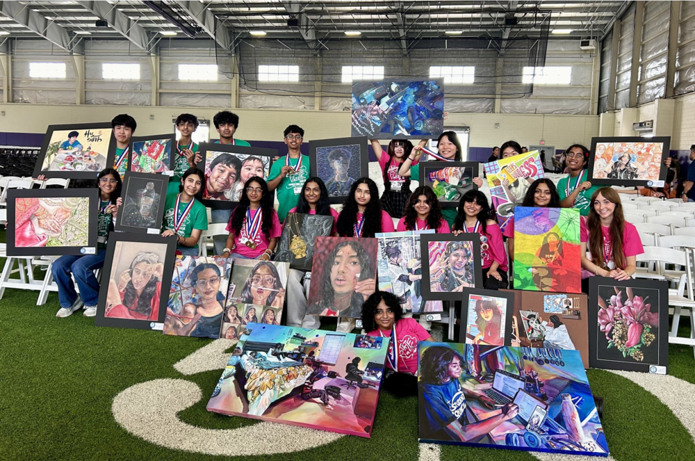 Large group photo taken inside an indoor fieldhouse or gymnasium. A group of students is arranged in rows, holding and displaying framed artworks of various styles and subjects, including detailed portraits, still lifes, interiors, and colorful abstract scenes. Many framed pieces are propped on the floor in front, while others are held at chest height. Several students wear medals around their necks, and many wear matching event T‑shirts. Rows of white folding chairs are visible in the background beneath a high metal ceiling, emphasizing the scale of the exhibition setting.