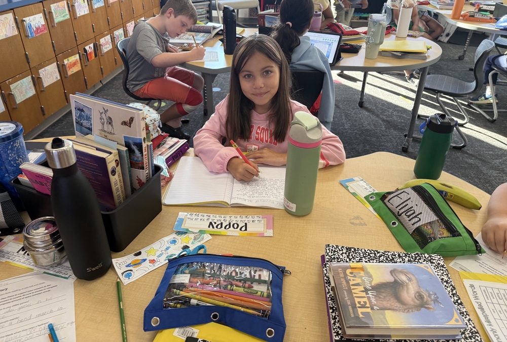 Student sits at a classroom table writing in an open notebook with books, pencils, folders and water bottles nearby. Other students work at tables in the background.