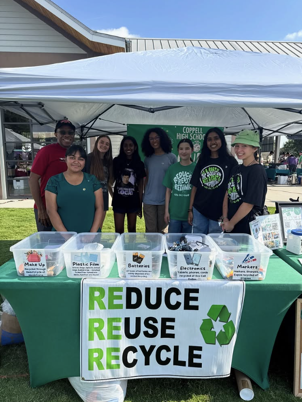 A group of Coppell High School Eco Club students stands behind a green table at an outdoor event under a white canopy. A sign on the front of the table reads “REDUCE REUSE RECYCLE” with a recycling symbol. Clear bins on the table are labeled for items such as makeup, plastic film, batteries, electronics, and markers. A green “Coppell High School” banner is displayed behind the group, with buildings and grass visible in the background.