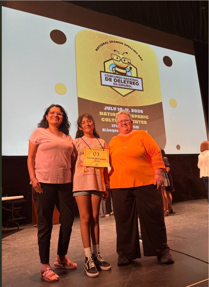 Three individuals stand on a stage during the National Spanish Spelling Bee (Concurso Nacional de Deletreo en Español). The person in the center holds a sign reading “03 Elizabeth Castillo, Texas.” Behind them, a large screen displays the National Spanish Spelling Bee logo and event information: “July 10-11, 2026, National Hispanic Cultural Center, Albuquerque, NM.” Additional participants are visible in the background on the stage.