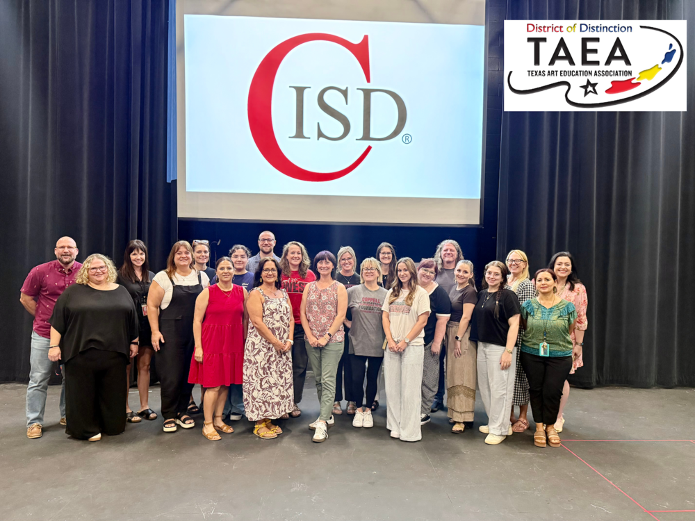 Group photo of approximately 20 Coppell ISD fine arts educators and staff standing on a stage in front of a large screen displaying the Coppell ISD logo. An additional graphic in the upper-right corner shows the Texas Art Education Association (TAEA) District of Distinction logo. The group is arranged in several rows against a backdrop of dark stage curtains. Attendees are wearing a mix of professional and casual attire. The stage floor and projection screen are clearly visible, and the photo appears to commemorate Coppell ISD’s recognition as a TAEA District of Distinction.