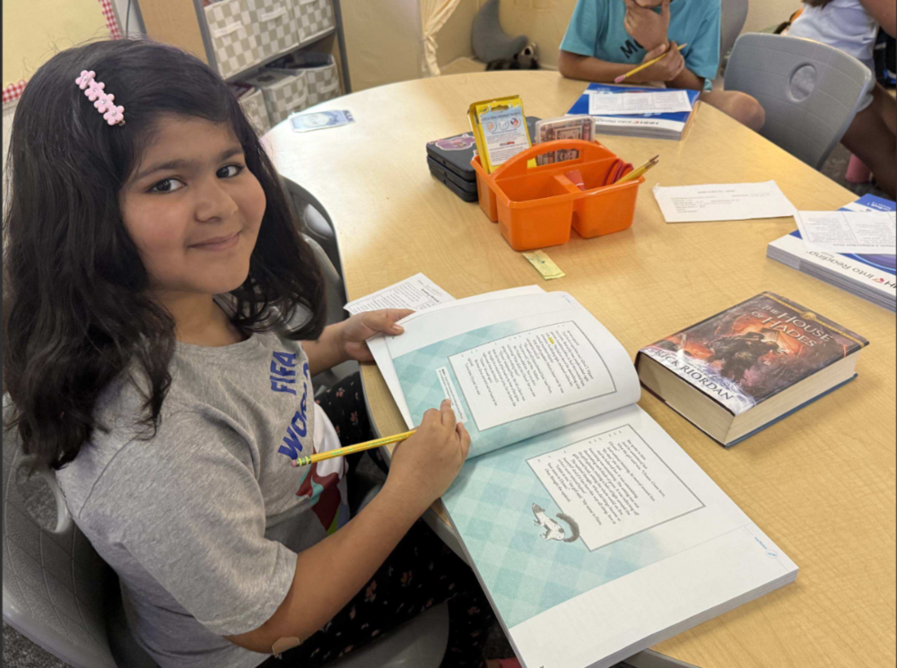 Photo of a student seated at a classroom table with an open book and pencil, smiling toward the camera while other students work nearby.