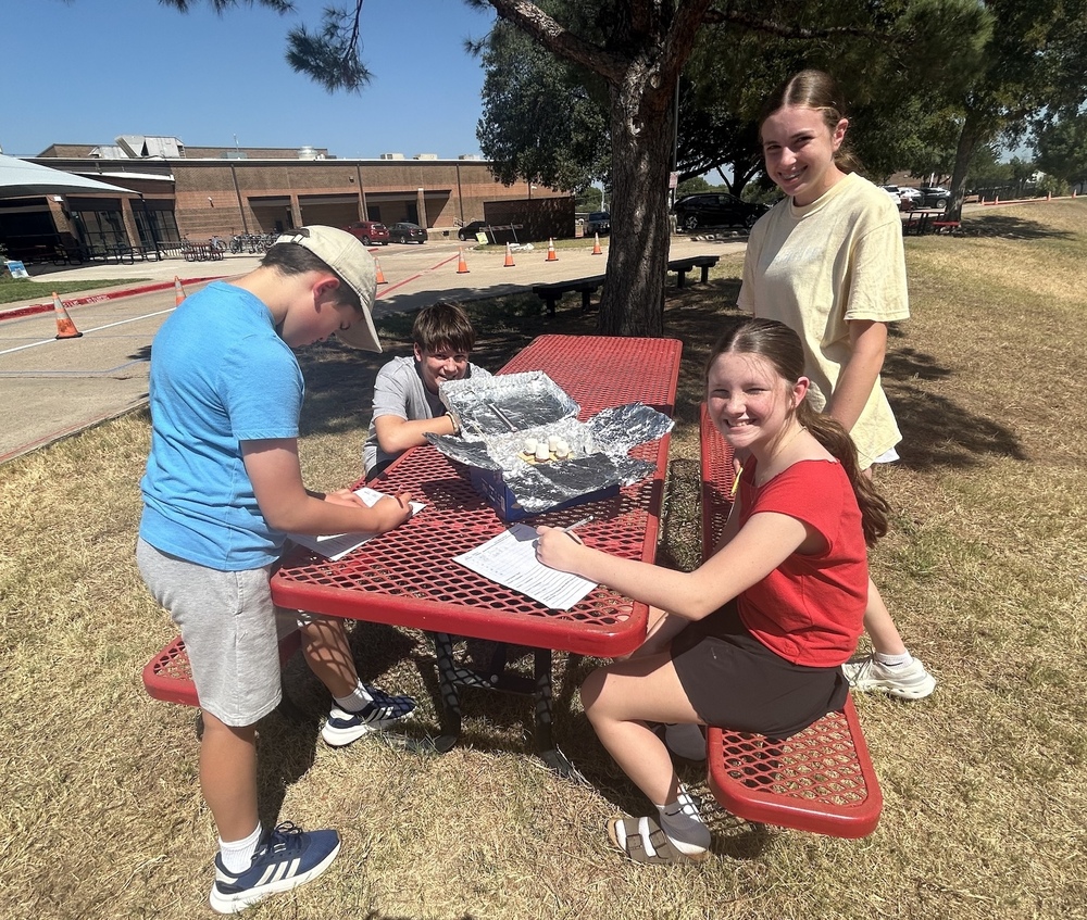 Students sit at an outdoor picnic table during a hands-on science activity, using notebooks and worksheets to record observations about foil-covered materials placed in the center of the tabl
