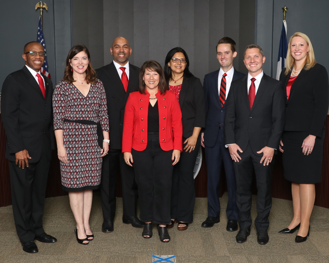 Group photo of eight Coppell ISD board of trustees and the superintendent, dressed in business attire, smiling together in front of a stage backdrop with American and Texas flags.