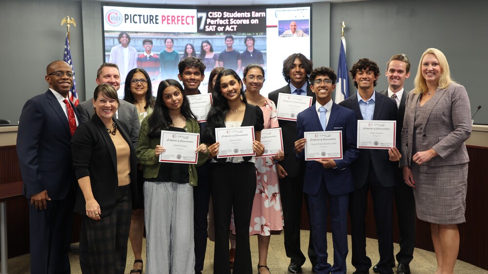 A group of Coppell ISD students and district leaders pose for a recognition photo in a boardroom. Several students in the front row hold certificates of commendation. A presentation screen behind the group displays the headline, “Picture Perfect: 7 CISD Students Earn Perfect Scores on SAT or ACT,” along with a photo of the recognized students. American and Texas flags stand on either side of the room. The group is gathered to celebrate students who achieved perfect scores on college entrance exams.