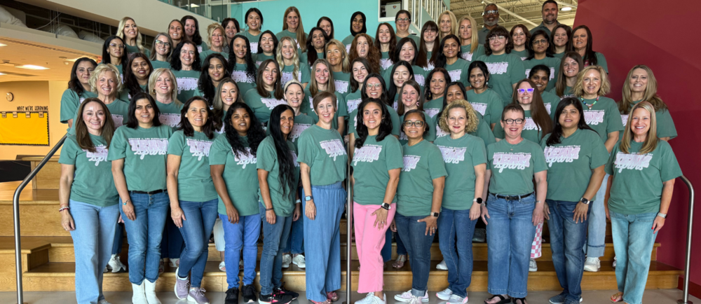 Richard J. Lee School staff pose together on indoor steps, wearing matching green T-shirts and jeans. Behind them, a bulletin board reads, “WHAT WE’RE LEARNING.