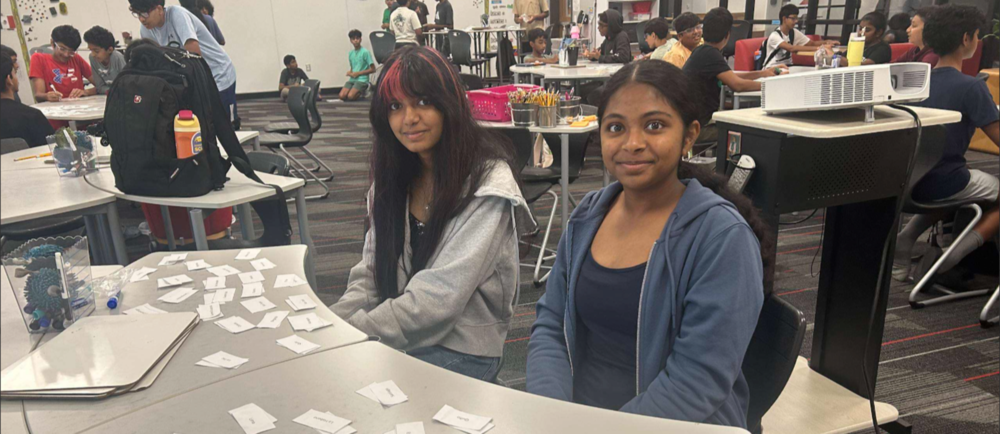 Two students sit at a table in a classroom while working with small paper cards, as other students collaborate at tables in the background.